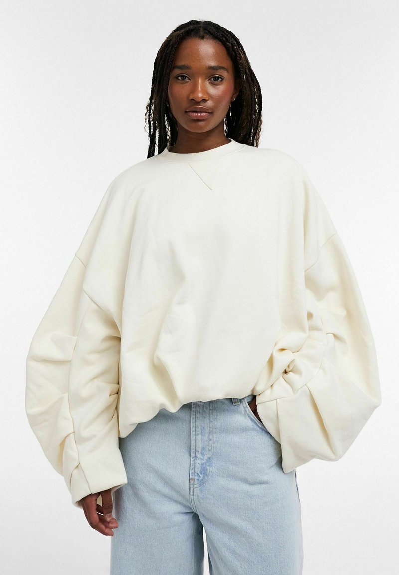 Femme aux longs cheveux tressés portant un sweat-shirt blanc oversize et un jean bleu clair, debout devant un fond blanc uni.