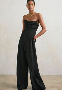 Svart satin jumpsuit utan axelremmar med en draperad bodice, vida byxor och sidofickor. Accenterad med ett guldhalsband. Slät textur.