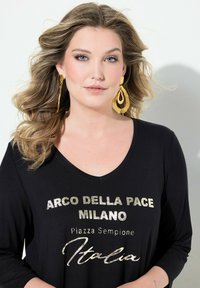 Zwarte lange mouwen top met metallic zilveren tekstprints met de tekst "ARCO DELLA PACE MILANO" en "Piazza Sempione Italia." Grote gouden statement oorbellen.