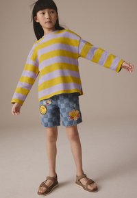 Haut à manches longues rayé violet et jaune associé à un short en denim à carreaux décoré de patchs smiley et fleurs. Sandales marron complètent la tenue.