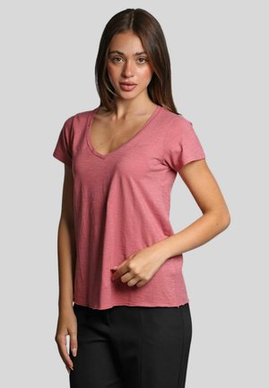Donna con lunghi capelli castani che indossa una maglietta rosa a scollo a V con maniche corte e pantaloni neri, in piedi di fronte a uno sfondo grigio chiaro uniforme.
