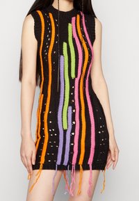 Abito in maglia nera con strisce multicolori (arancio, rosa, verde, viola) e dettagli intagliati; presenta frange sull'orlo e un colletto alto.