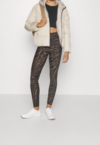 Puffjakke i lyse beige, parret med sorte mønstrede leggings med svulmende linjer og hvirvler, samt hvide sportssko.