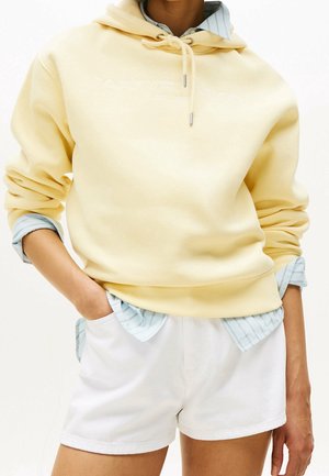 Persona con una sudadera con capucha amarillo claro sobre una camisa azul a rayas y pantalones cortos blancos, con las manos en los bolsillos, contra un fondo liso.