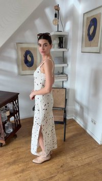 Γυναίκα με λευκό αμάνικο φόρεμα με καφέ βούλες και μπεζ μποτάκια στέκεται σε ένα δωμάτιο με ξύλινο πάτωμα και κορνιζαρισμένο μπλε έργο τέχνης.