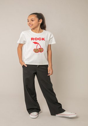 Fille aux cheveux foncés attachés en queue de cheval, portant un t-shirt blanc "ROCK" avec des cerises imprimées en léopard, un pantalon noir et des baskets blanches, debout les mains dans les poches.