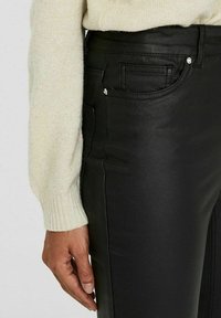 Svarta fuskläderbyxor med slim fit, med framfickor och subtila sömdetaljer. En ljus krämfärgad tröja syns vid handleden.