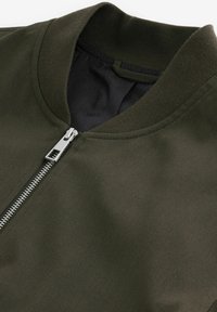 Olivgrüne Bomberjacke aus glattem Stoff, mit geripptem Kragen und silbernem Reißverschluss. Das Innenfutter besteht aus schwarzem Mesh.