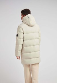 Uomo che indossa un giubbotto imbottito color crema con cappuccio, in piedi e girato di spalle su uno sfondo chiaro e semplice.