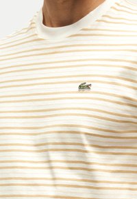 Άνδρας που φοράει άσπρο και μπεζ ριγέ πουκάμισο με πράσινο λογότυπο κροκοδείλου στο στήθος.