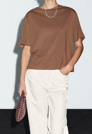 Person iført en brun løs T-shirt, off-white bukser, sølvkædehalskæde, holder en mørk vævet clutch i den ene hånd med den anden hånd i lommen.