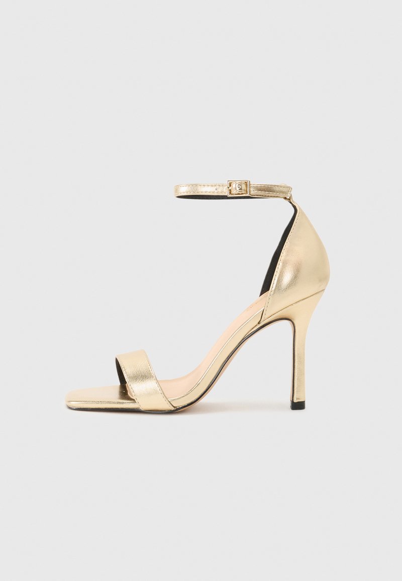 Zlati metalni visokodebelni sandal z ozkim gležnjam, odprtim nosom in elegantnim dizajnom. Ima gladko teksturo in minimalistične detajle.