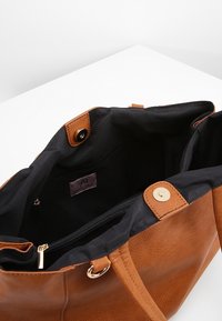 Ruskea nahkainen olkalaukku, jossa on kultaiset yksityiskohdat. Laukussa on musta kangaspinnoite, sisäinen vetoketjupussi ja päällinen napillinen suljenta.