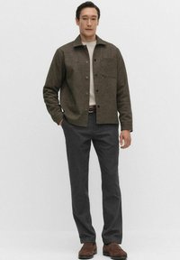 Bruine getextureerde jas met kraag, voorzak en knoopsluiting; gedragen over een lichte trui en donkergrijze op maat gemaakte broek met bruine schoenen.