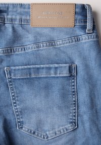 Vaaleansiniset farkut, joissa on tekstuuripinta, yhdellä takataskulla ja ruskealla nahkahihnalapulla, jossa on kuvitettu teksti.