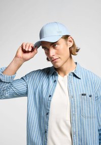 Jasnoniebieska czapka z zakrzywionym daszkiem i wytłoczonym logo, noszona z niebieską w paski koszulą z guzikami na białym t-shircie.