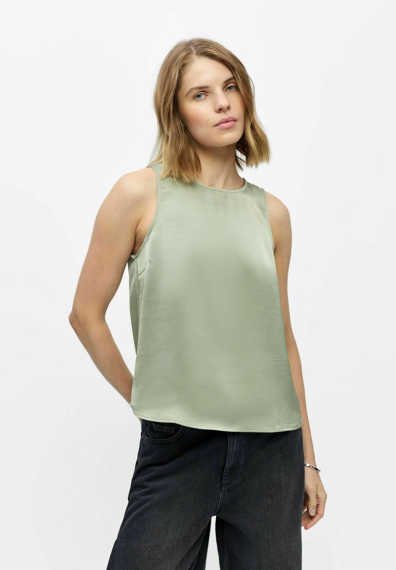 Femme aux cheveux blonds mi-longs, portant un haut sans manches vert clair et un jean foncé, debout devant un fond blanc uni.