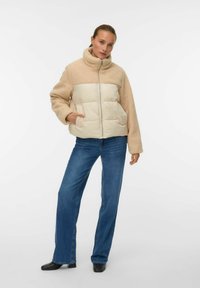 Sieviete, tērpusies bēša krāsas flīsa un šūta jaka ar priekšējām kabatām, apvienota ar zilām džinsu biksēm un melniem kvadrātveida purngala apaviem, stāv baltas sienas priekšā.