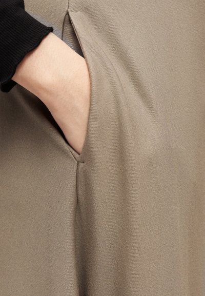 Χέρι σε τσέπη μπεζ παντελονιού, φορώντας μαύρο μακρυμάνικο τοπ, που δείχνει την υφή του υφάσματος και το ράψιμο στην άκρη της τσέπης.