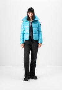 Vrouw met steil, donker haar in een felblauwe pufferjacket, zwarte jeans en zwarte plateau schoenen tegen een witte achtergrond.