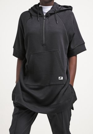 Person iført en oversized sort kortærmet hættetrøje med lynlås foran, stor kængurulomme, Nike-logo patch og sorte bukser.