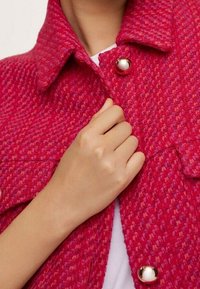 Czerwona i różowa teksturowana kurtka z kołnierzem, zapięciem na guzik oraz widoczną ręką trzymającą materiał. Wyraźny wzór tkany.
