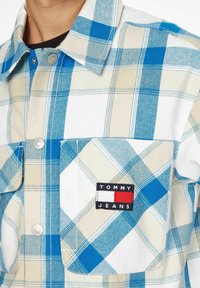 Blå och tan rutigt flanellskjorta med krage som knäpps, som har en bröstficka och en märkespatch med "TOMMY JEANS."