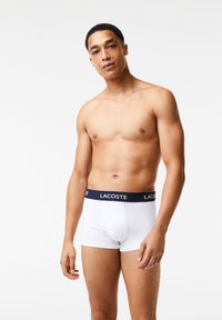 Fehér boxer alsónadrág sötétkék derékszíjjal, amelyen a "LACOSTE" logó található. Sima anyag, testhezálló fazon és combközép hosszal.