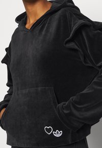 Svart velour hoodie med en frampå ficka, volangärmar och vita broderade hjärtan och logotypdetaljer. Mjuk textur och avslappnad passform.