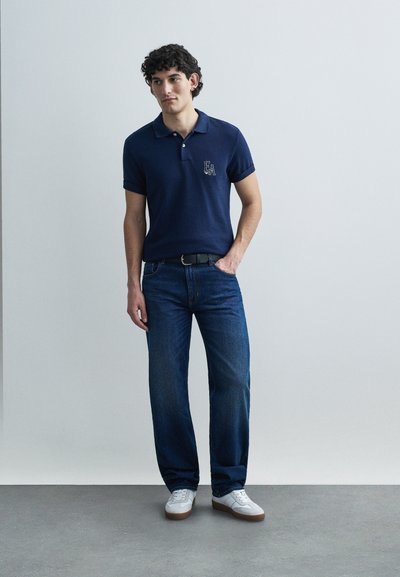 Μπλε ναυτικό πόλο με κοντά μανίκια και ριγωτή υφή, συνδυασμένο με τζιν παντελόνι ίσιου ποδιού και λευκά αθλητικά παπούτσια. Διακριτικό λογότυπο στο πόλο.