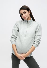 Šviesiai pilkas džemperis su aukštu apykakliu, pusiau užtrauktuku ir džiuto rankogaliais. Minkšta tekstūra ir laisvas siluetas, derinamas su tamsiai pilkomis kelnėmis.