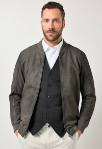 Man met korte baard, draagt een donkergrijze bomberjack met rits over een zwart knoopvest en wit overhemd, handen in de zakken van lichte broek.