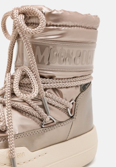 Beige, vadderad månskos med en glansig yta, tjocka snören, metallögon, och en texturerad gummisula med logotryck.