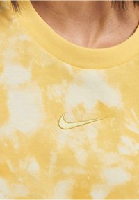 Geltonas tinklinio marškinėlis su apykakle, kuriame yra mažas auksinis Nike logotipas ant krūtinės. Lygus audinio tekstūros su kasdiene dizainu.