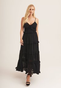 Black maxi dress with ruffle detailing, spaghetti straps, and tiered layers. Lightweight fabric with a flowing silhouette. Black high heels.