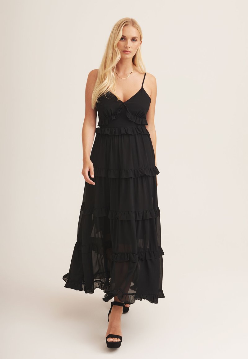 Black maxi dress with ruffle detailing, spaghetti straps, and tiered layers. Lightweight fabric with a flowing silhouette. Black high heels.