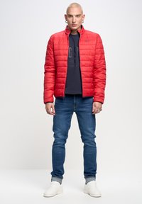 Sarkans kvilts jaka ar stāvu apkakli, uzvilkta virs tumši zilā krekla ar garām piedurknēm, apvienota ar ziliem džinsiem un baltiem sporta apaviem. Piegriezums ir izsmalcināts, virsma gluda.