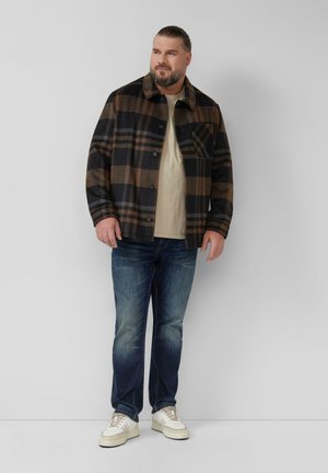 Man met baard in een bruine en zwarte geruite jas, beige shirt, blauwe spijkerbroek en witte sneakers, staande tegen een effen witte muur.