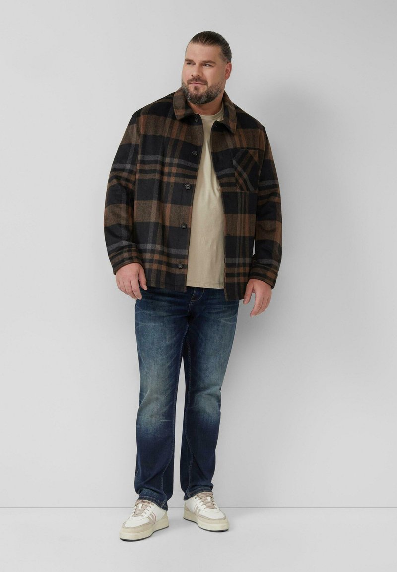 Man met baard in een bruine en zwarte geruite jas, beige shirt, blauwe spijkerbroek en witte sneakers, staande tegen een effen witte muur.