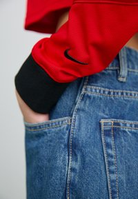 Piros, légáteresztő anyagú, hosszú ujjú póló fekete mandzsettával, fekete Nike logóval. Kék farmernadrággal párosítva, amelyen a zsebek és a varrás részletei láthatók.