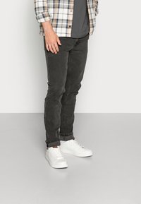 Vaqueros de mezclilla negros con corte slim y puños enrollados, combinados con zapatillas blancas. La camisa es de cuadros en tonos crema, azul y marrón.