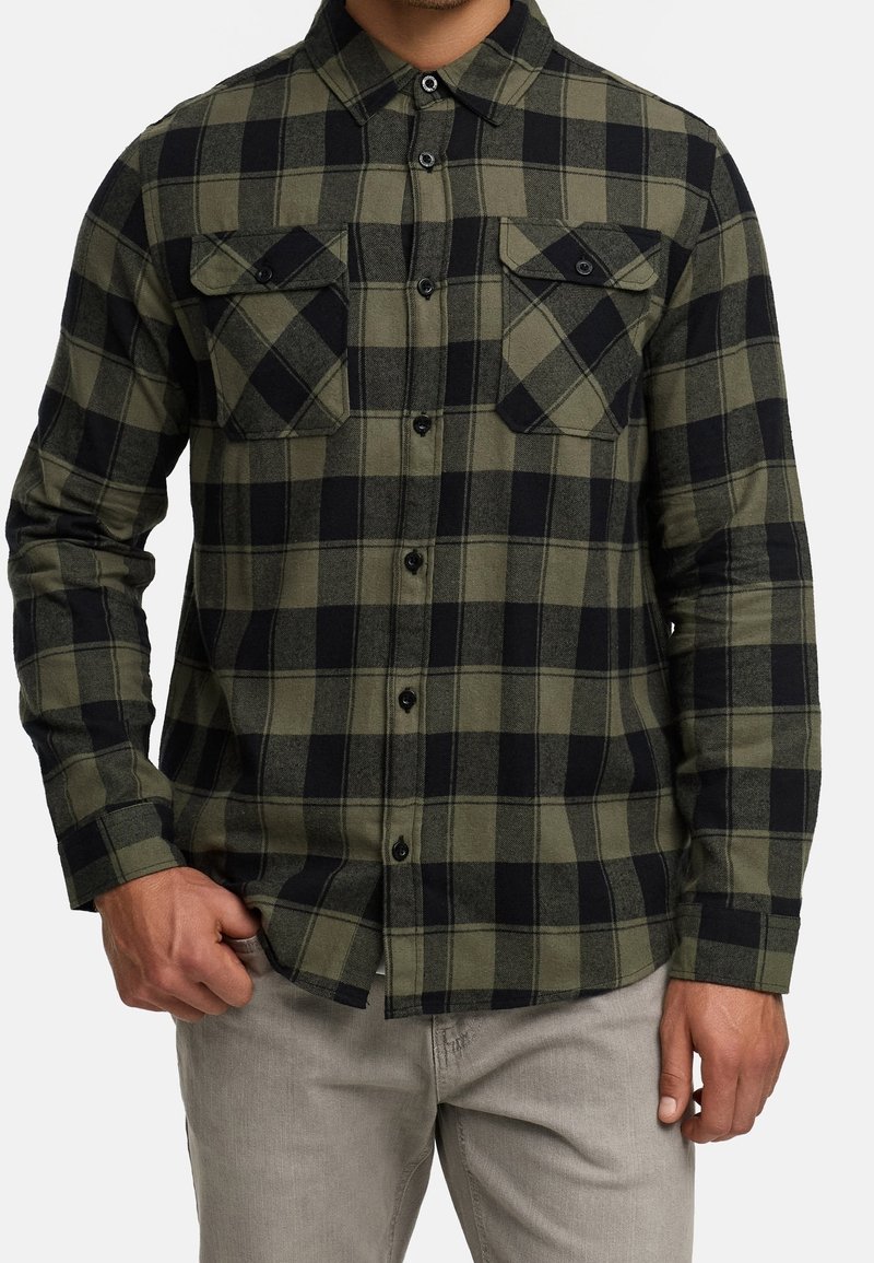 Homme portant une chemise en flanelle à carreaux vert olive et noir à manches longues, boutonnée, avec deux poches poitrine et un pantalon gris clair.