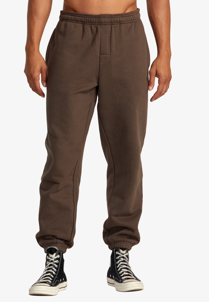 Braune Baumwoll-Sweatpants mit elastischem Bund, seitlichen Taschen und gerippten Bündchen, kombiniert mit schwarzen High-Top-Sneakern.