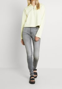 Suéter corto de color amarillo claro con cuello de cremallera, combinado con pantalones ajustados gris y sandalias negras con detalle de hebilla.