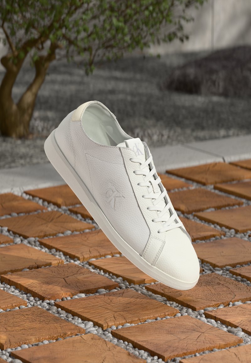 Witte leren Calvin Klein sneaker met veters die rusten boven houten vierkante tegels en kleine grijze stenen buitenshuis.