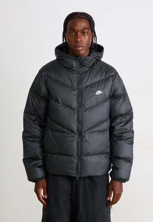 Black puffer jacket with a zip closure and hood. Features a chevron quilted design and a small white Nike logo on the chest.
