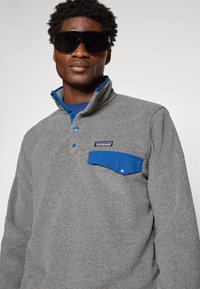 Man in een oversized zwarte zonnebril en een grijze Patagonia fleece met een blauwe zakflap en kraagaccenten.