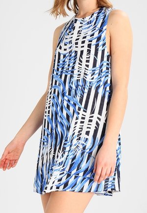 Robe sans manches à rayures verticales noires et blanches avec des motifs de feuilles bleues portée par une femme sur un fond uni.