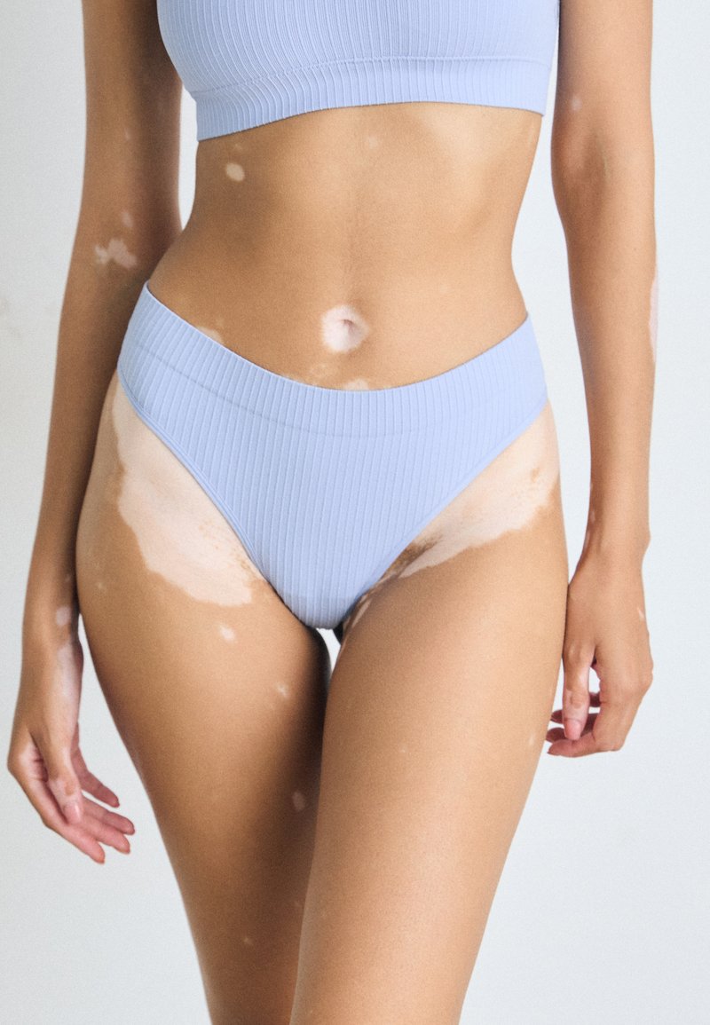 Femme atteinte de vitiligo portant des sous-vêtements en rib et un crop top bleu clair, se tenant avec les bras détendus devant un fond uni.