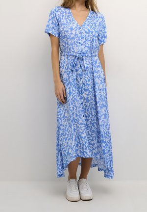 Femme portant une robe midi boutonnée à fleurs bleues avec des manches courtes et une ceinture à la taille, associée à des baskets blanches, debout contre un mur gris.